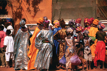 ​​​​​​​Fulani women attending the Ouahigouya regional fair. Colorfully dressed women with headwraps are seen from behind against an orange wall. 