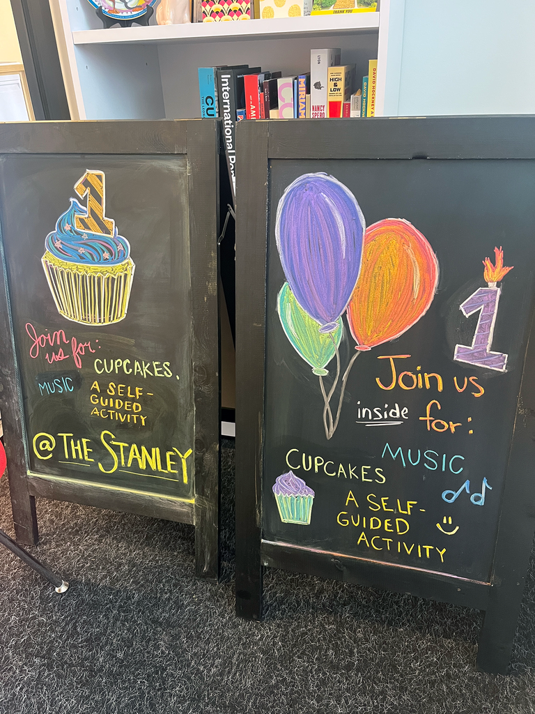 A photo of two chalk sandwich board signs in an office. One sign has a cupcake drawn on it, the other has multicolored balloons and a "1" birthday candle. Both advertise the HBD, SMA birthday party event.