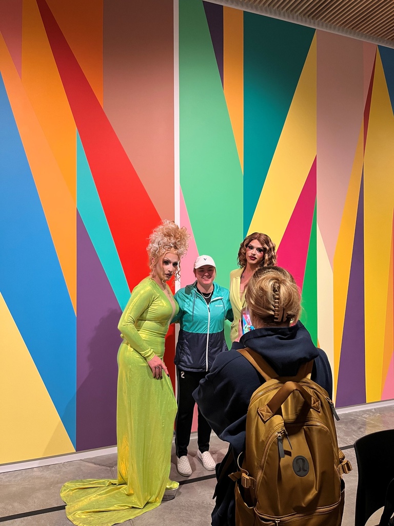 shown is a student standing in between two drag queens against the lobby mural 
