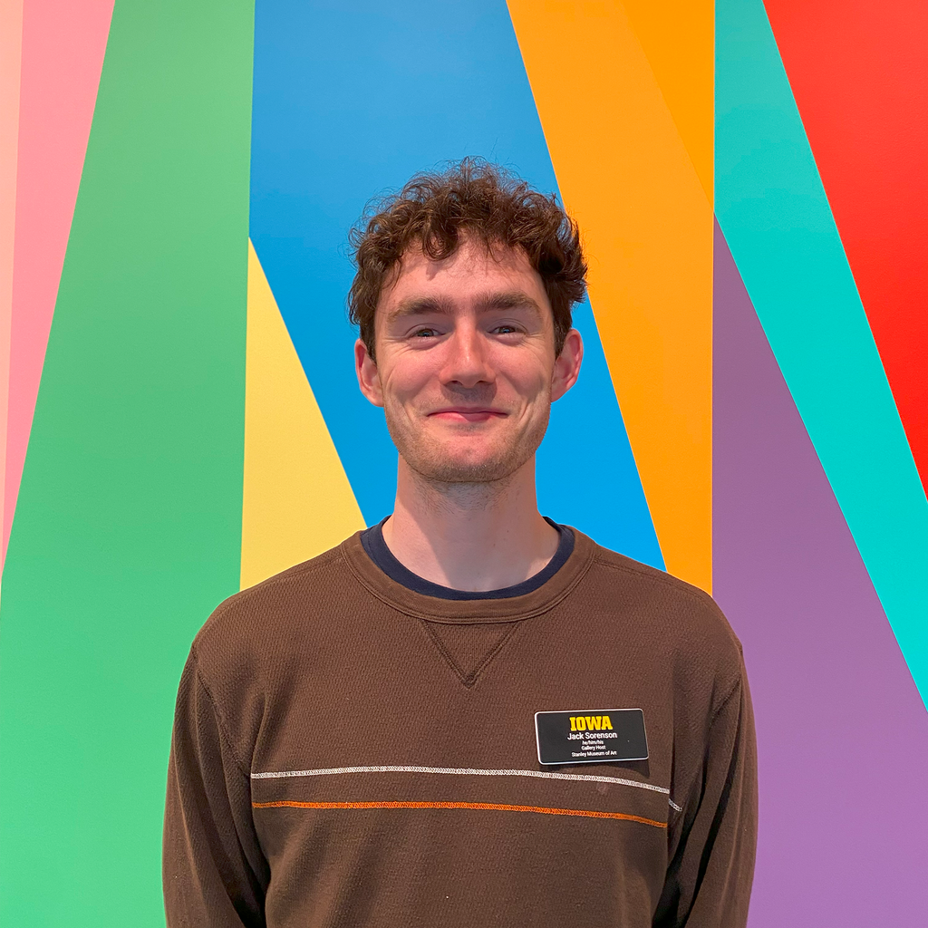 A photo of gallery host Jack Sorenson. He stands, smiling, with his hands behind his back in front of the colorful lobby mural at the Stanley Museum of Art.