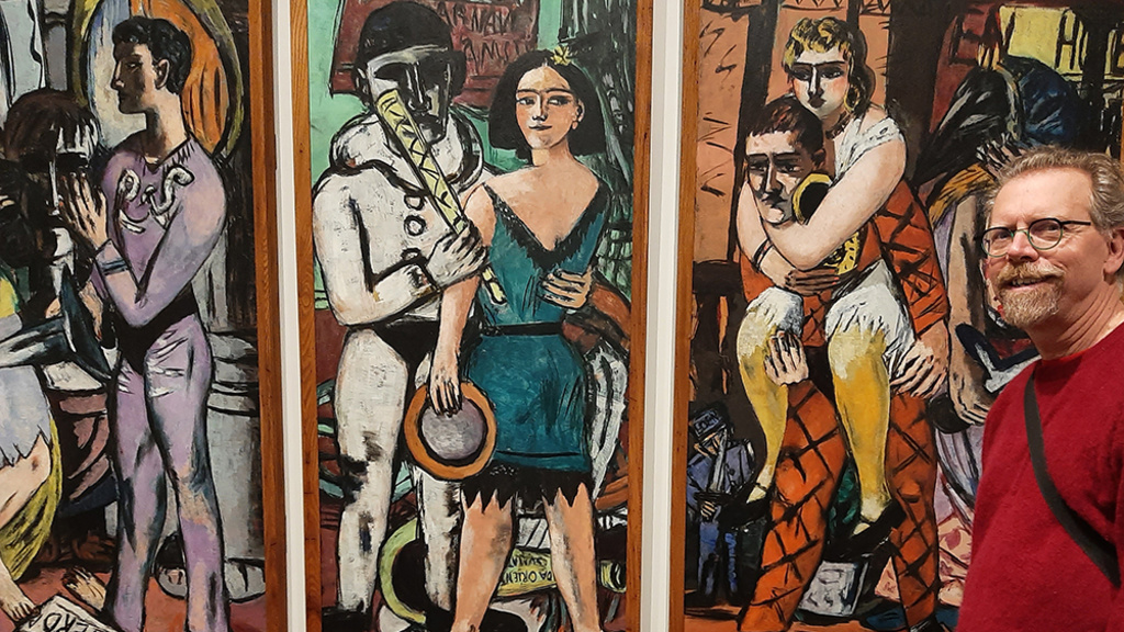 A man with beard and glasses stands in front of a three-panel painting. L: Person in yellow dress stands, one leg crosses the other, plays a flute next to figure in purple holding a knife. Behind is large sculpted head, partly in shadow. Cntr: Person in black and blue dress clutches circular object, holds long cylindrical object like a horn, stands close to figure in a black and white clown costume.  R: Tired harlequin clown carries a jewelry clad person piggyback.