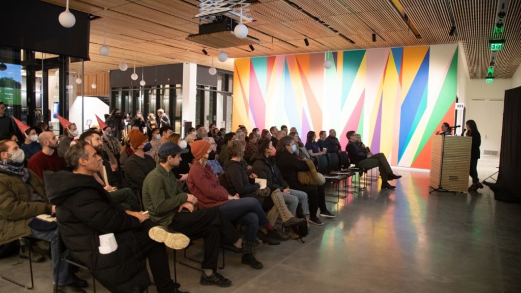 A seated audience listens to a speaker, there is a brightly colored, geometric wall mural behind them.