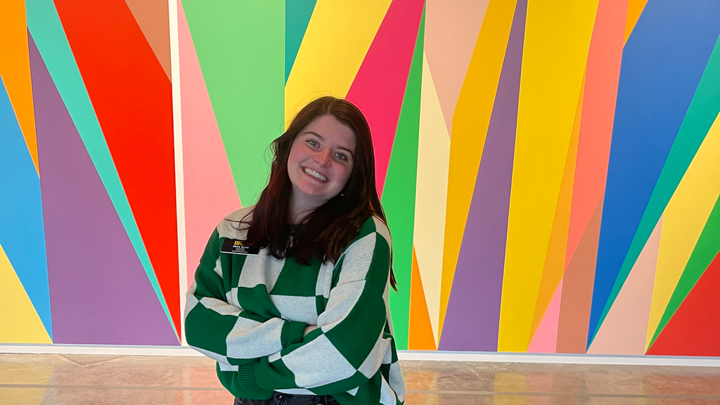A photo of Alexis Belme: she is standing in front of the mural in the Stanley Museum of Art lobby, smiling, her arms crossed.