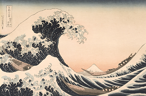 Woodblock print of a large dark blue wave with frothing white caps. The crest rises from the left and reaches out over a mountain in the middle which appears small and distant. Three long boats with people in grey robes ride the wave.