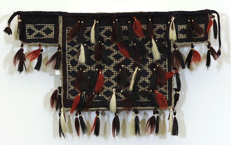A square woolen bag with two small rectangles extending from the top on either side. It is black and is woven with a motif of cream colored diamonds, and is covered in many goat hair tassels in cream, black, and red.