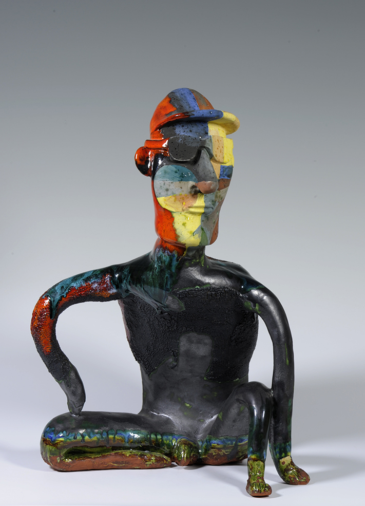 Seated male nude figure, leaning forward with arms arching away from the torso. Bright blocks of color cover the head, sunglasses, and a baseball cap, and paint drips down the right arm and in bands along the bottom portion of the sculpture.