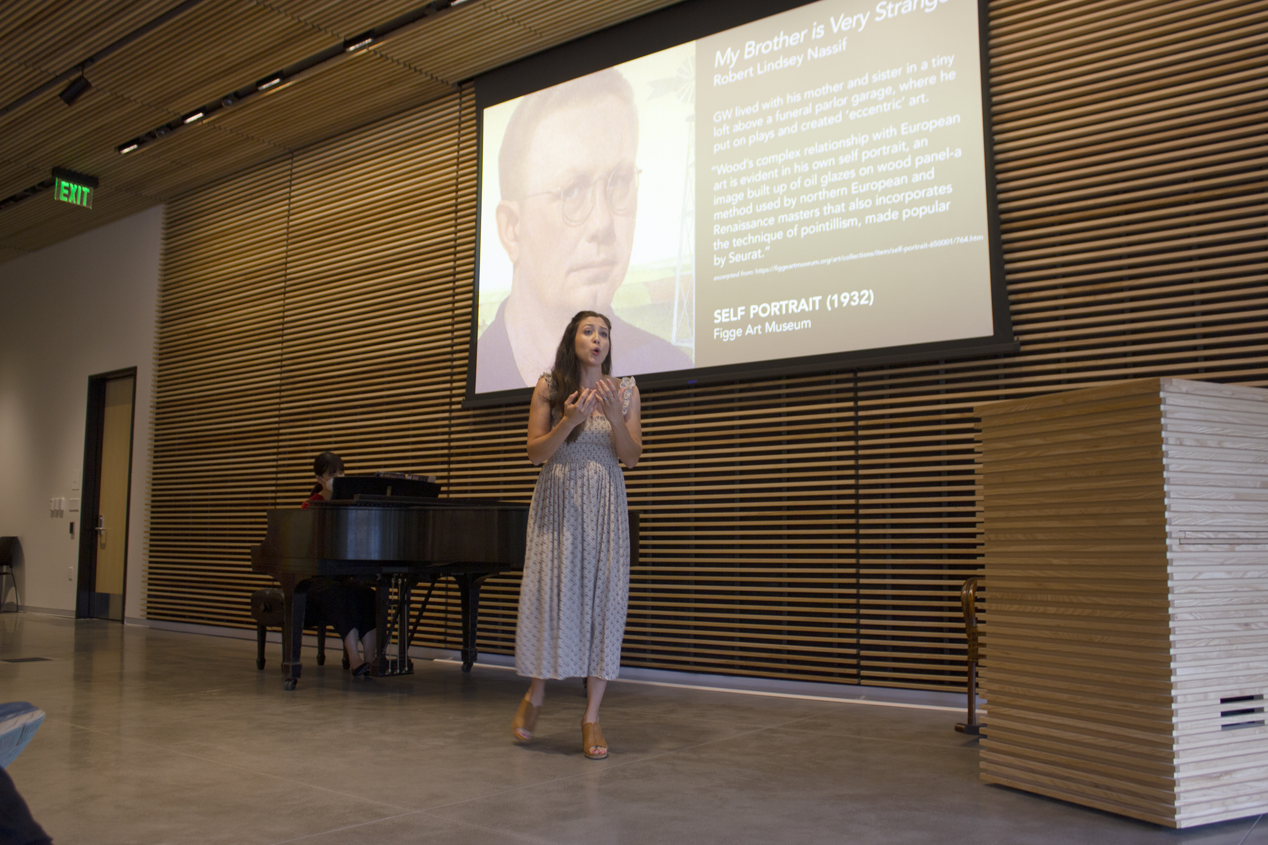 Female opera singer stands before a grand piano and a screen with a portrait of Grant Wood.