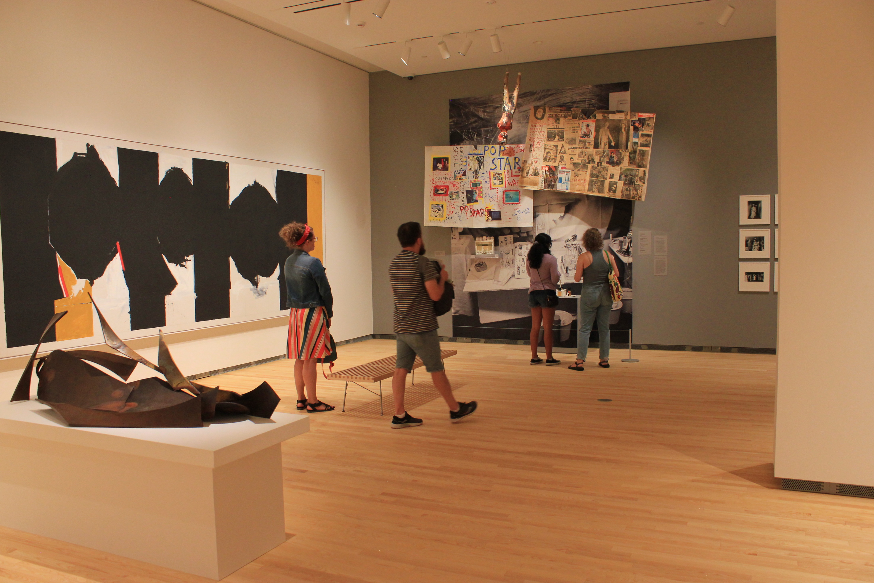 A group of visitors in an art gallery. There is a low-profile sculpture on a table in the foreground, a large, wide painting (with blacks, yellows and white background) on the left and an installation of hanging and standing works in the background