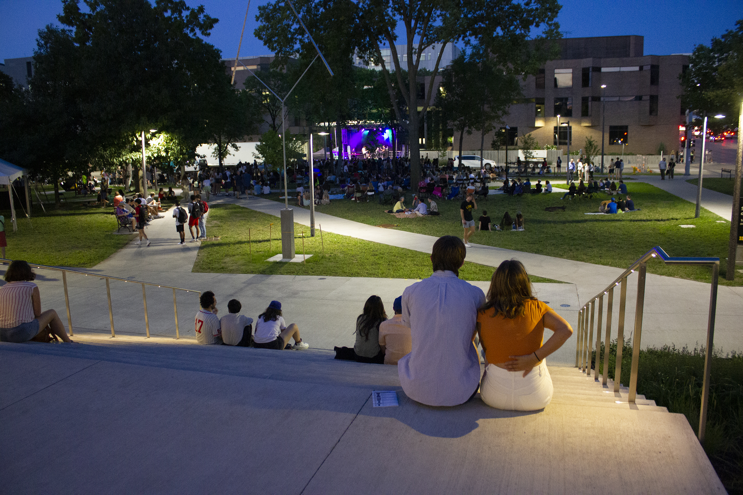 A man and woman sit at the top of a staircase look out over a park filled with people. There is a stage with a band in the distance