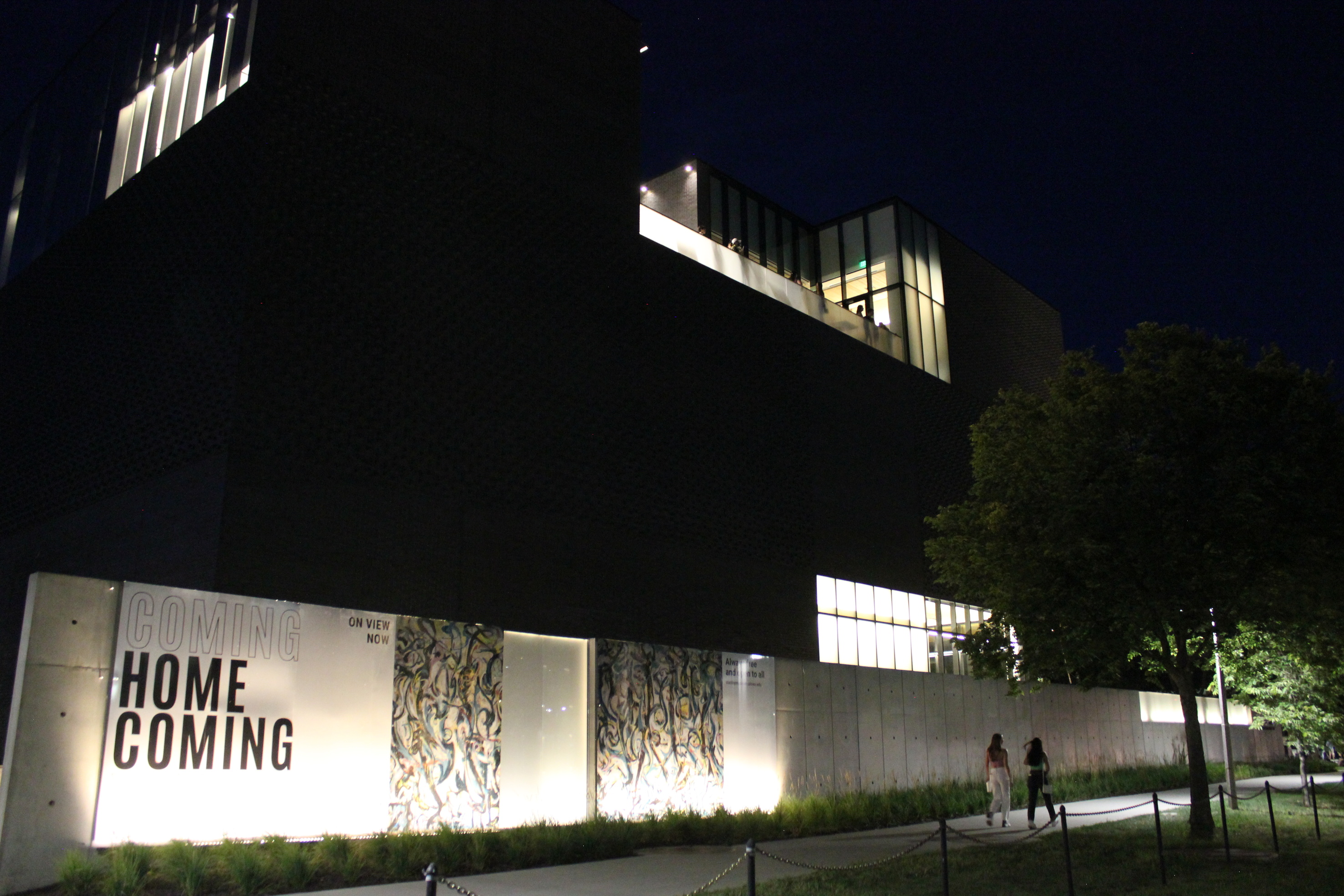 Exterior of museum building at night. A large sign with "Homecoming" is installed on a wall in the foreground and lit from below