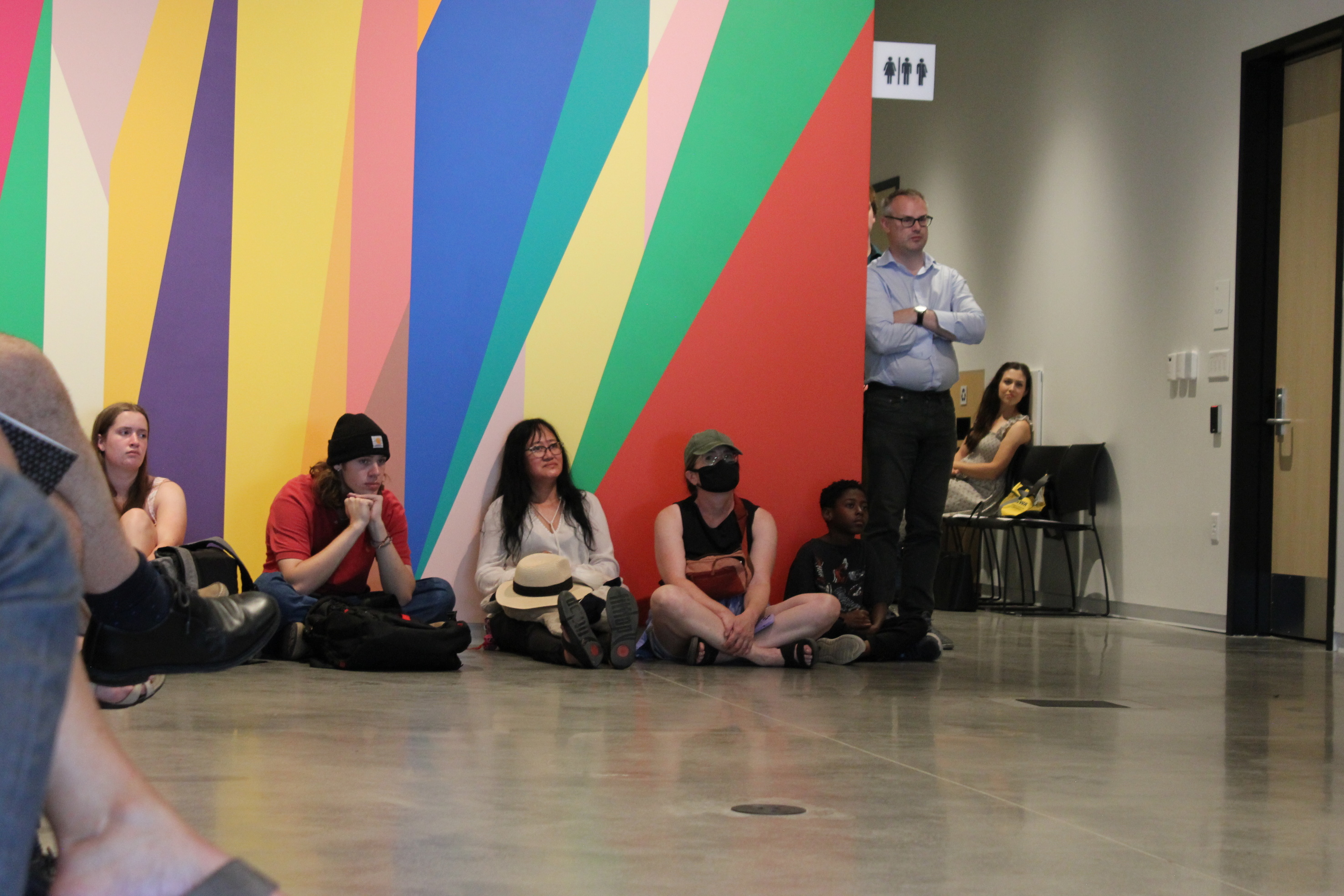 Visitor sitting on the floor to watch a performance in the lobby of building. There is a brightly-colored, geometric wall mural behind them