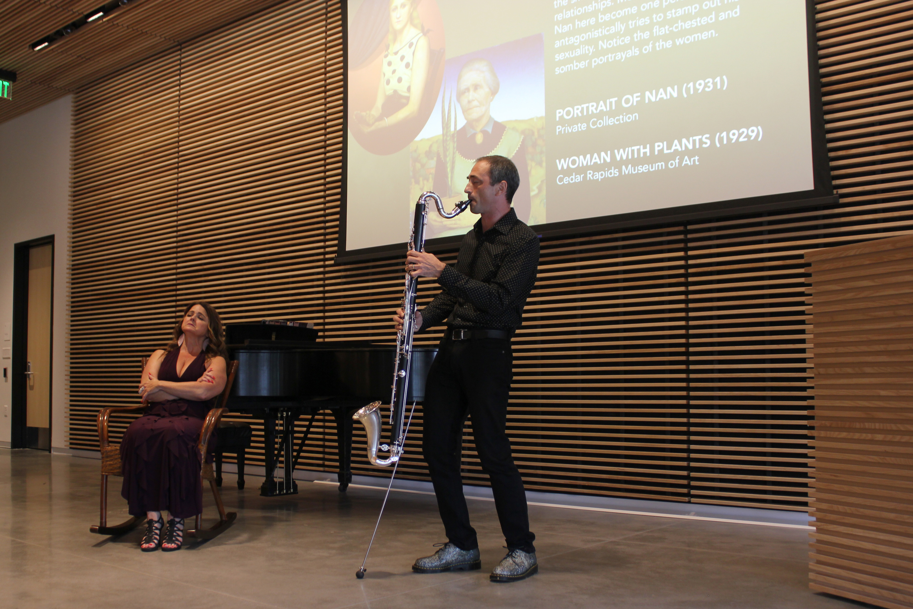 At left a woman in a dark purple dress sits in a rocking chair. At right a man stands playing a bass clarinet. 