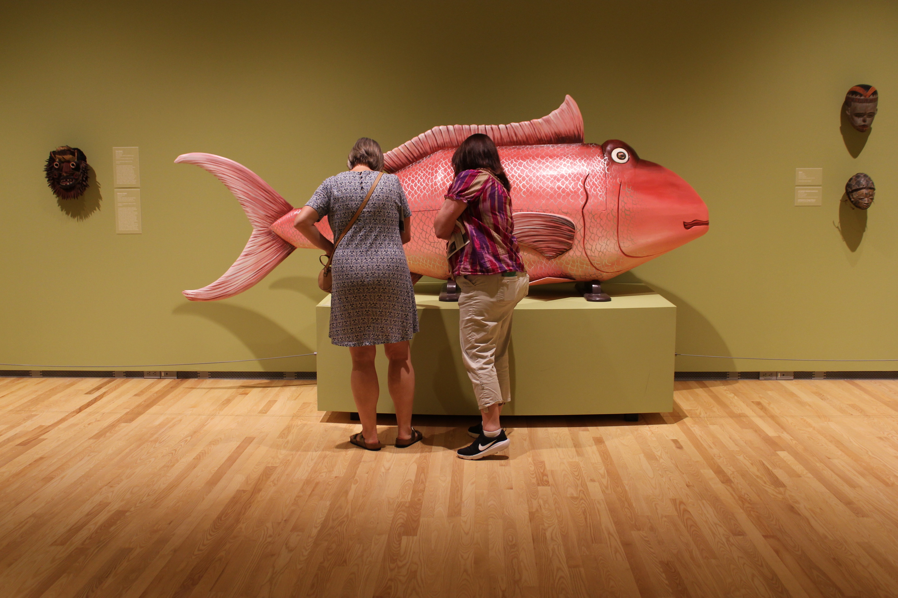 Two women stand in front of a large, pink fish sculpture.