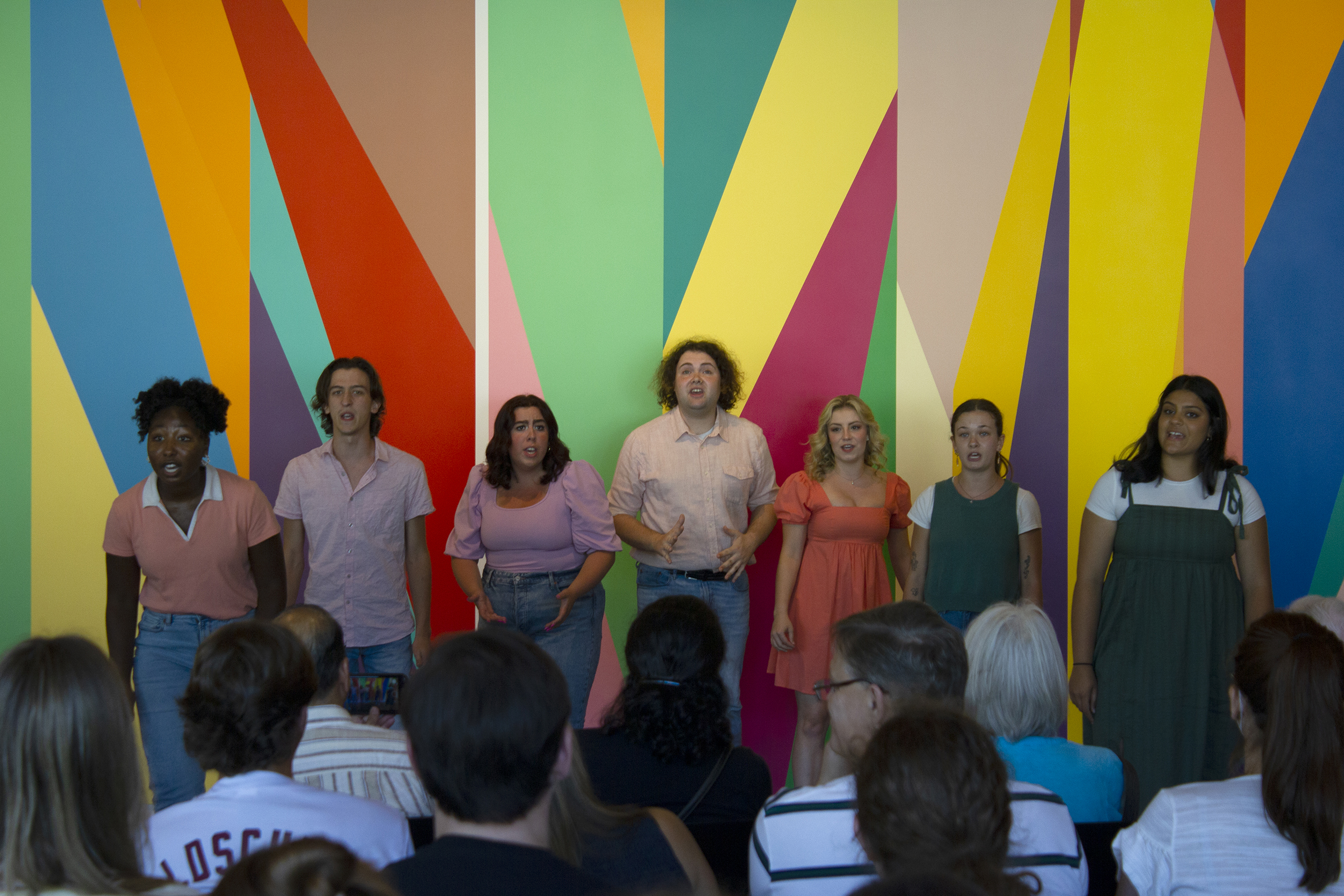 A line of musical theater students sing to a seated audience in front of a brightly colored, geometric wall mural