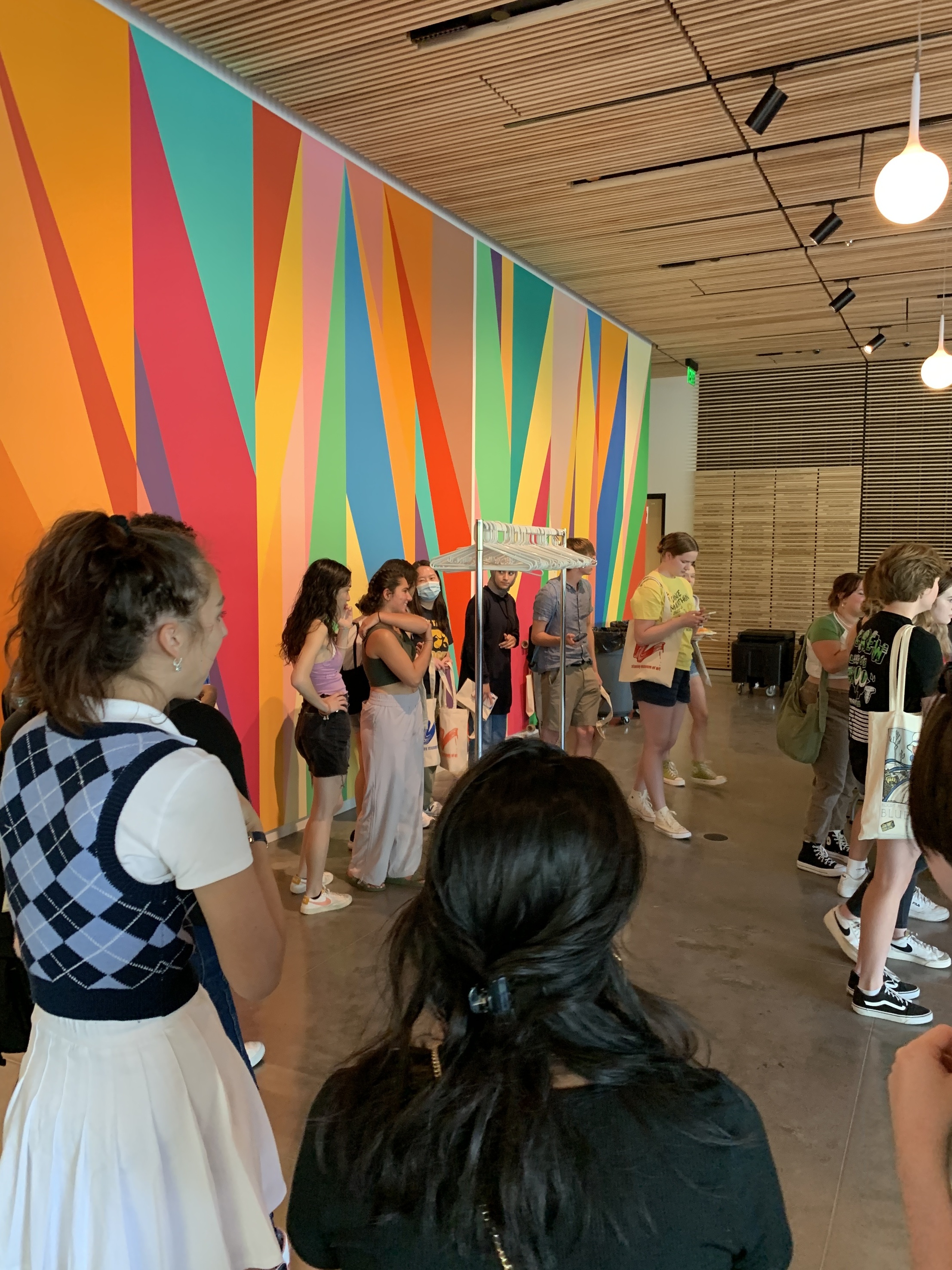Group of students select tote bags from a stand in front of a brightly colored wall mural