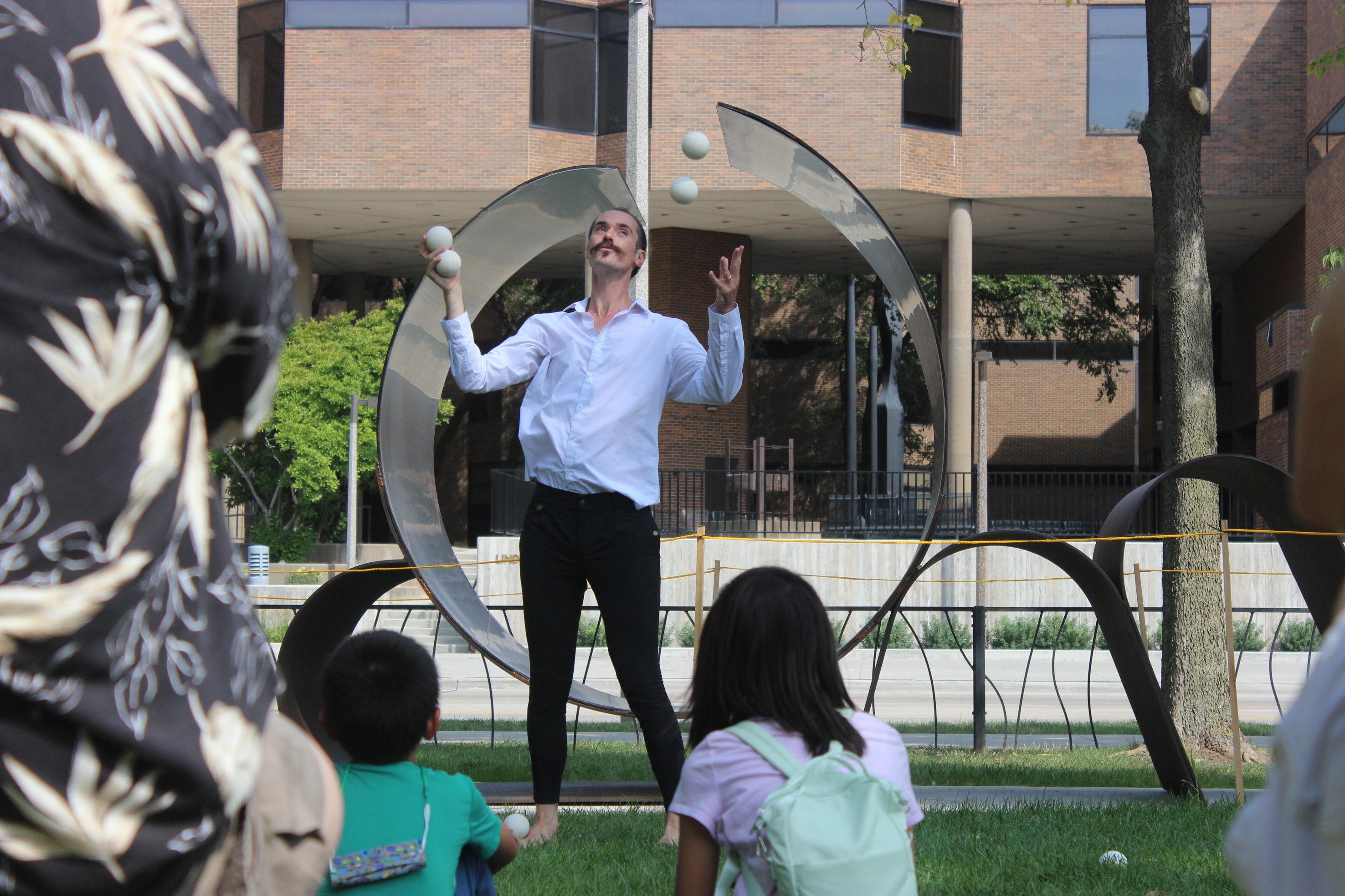 A man in a white shirt and dark pants juggles white balls in front of an outdoor sculpture