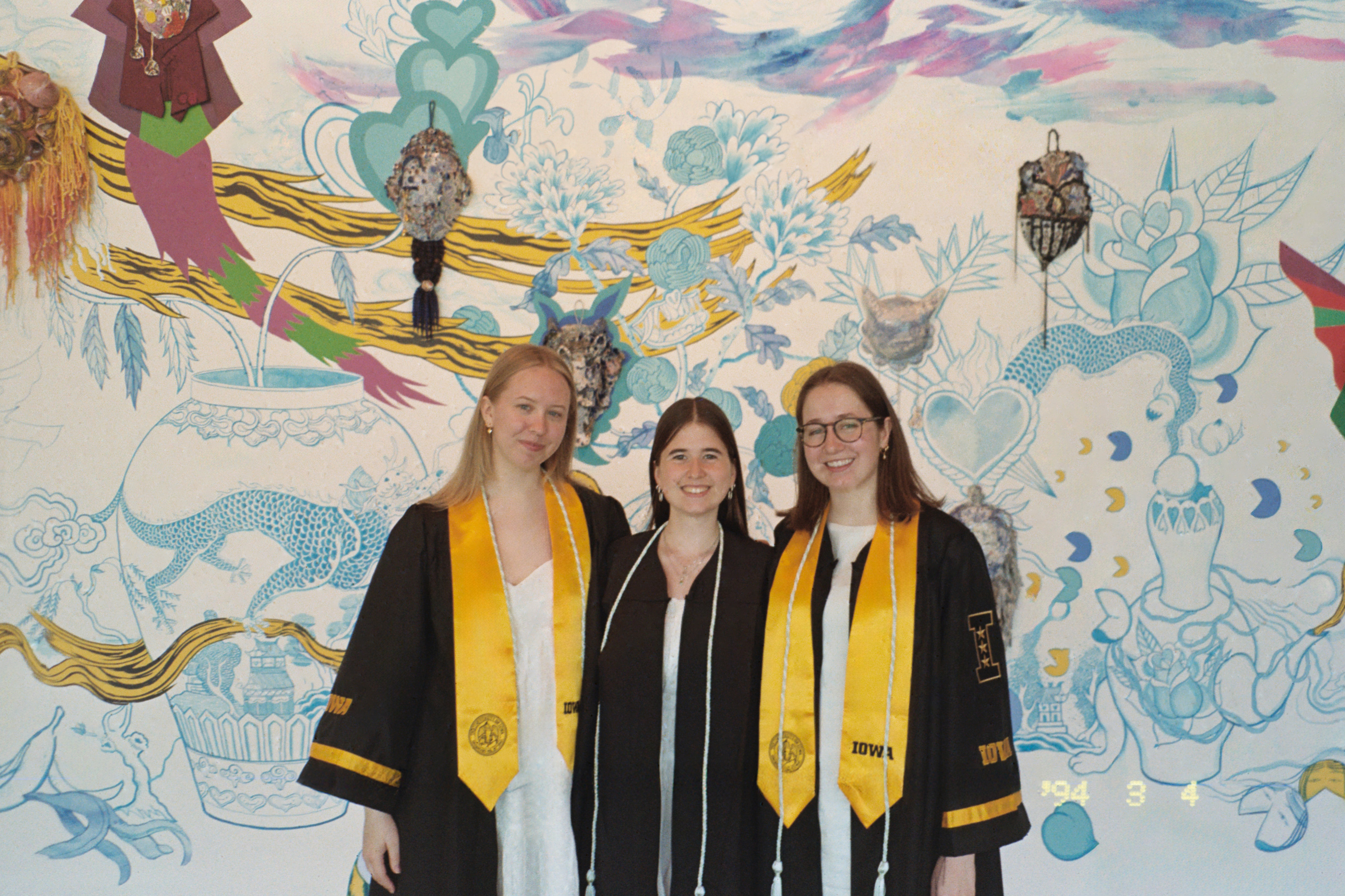 Student employees Josie Duccini, Annelies Knight, Abbie McLaren pose together in their graduation robes front of the Jiha Moon mural in the Stanley Museum of Art lobby.