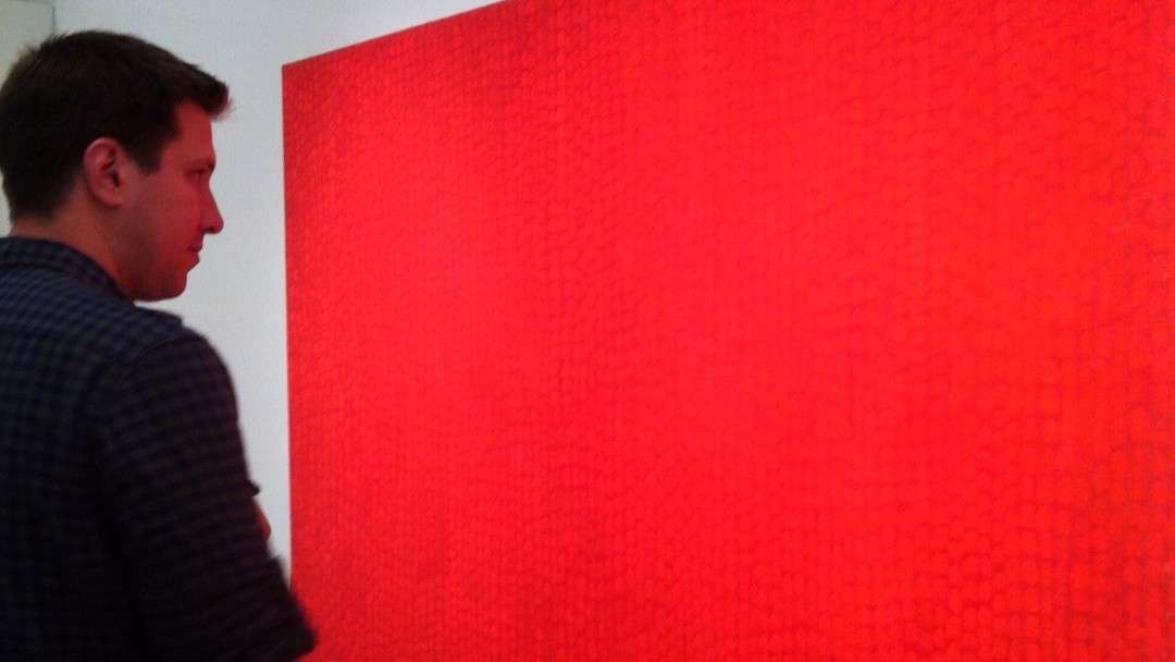 A man with short dark hair, wearing a dark shirt looks at Steve Erickson's monumental red painting, "Earth" 