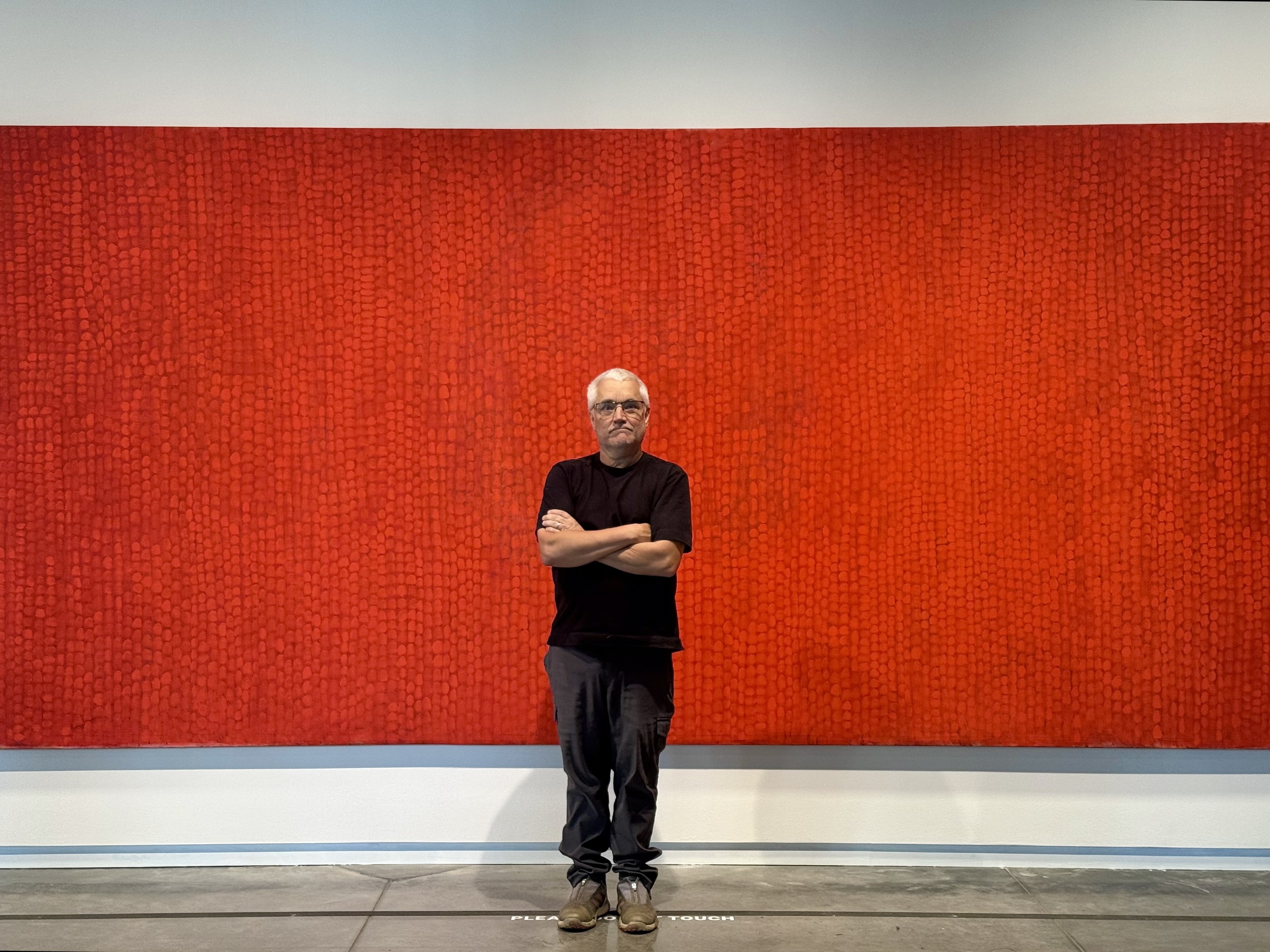 Artist Steve Erickson poses wearing a dark shirt and trousers, in front of his red canvas painting, "Earth," on view in the Stanley Museum of Art lobby