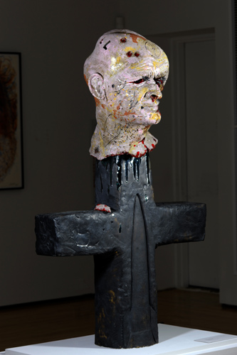 Sculpture of a corpse-like head (with words such as “slaughter” and “kill” stamped on the back)  bleeding on top of a black cross, on which there is a relief in the shape of a nuclear warhead.
