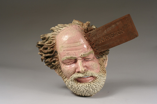 Ceramic head of bald man with white beard. There is a brick slamming into the side of his head. The brick has the word “Arneson” carved into it.