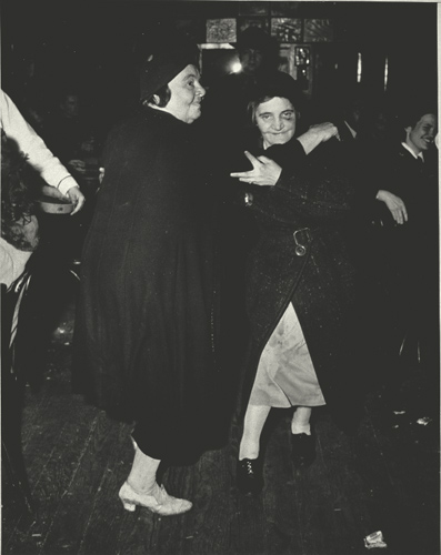 two women wearing long, dark coats dancing together
