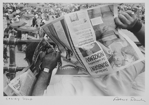 man holding an open newspaper over his head while he looks down at a beach crowded with people