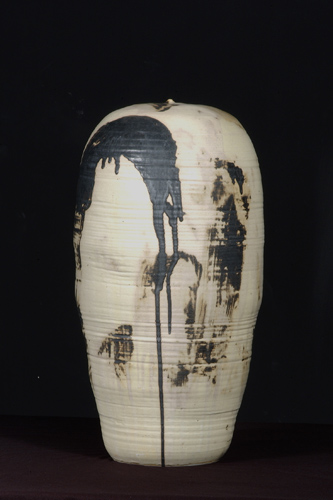 Tall oval-shaped stoneward object, glazed in a cream color with areas of dark brown that are splattered/dripping and smeared.