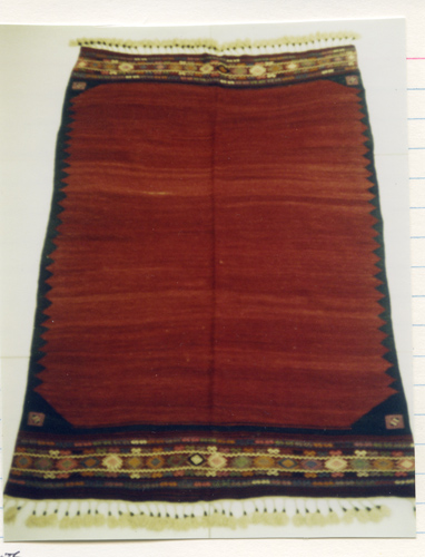 A large woolen rug which is primarily a deep red, and has detailed  woven designs in other muted colors as well as a row of cream colored tassels on either end.