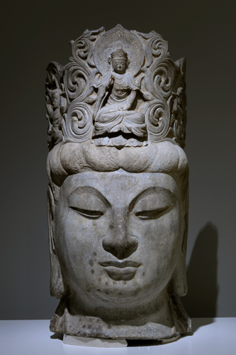 Carved head with tranquil face, eyes looking downward, and an elaborate headpiece.