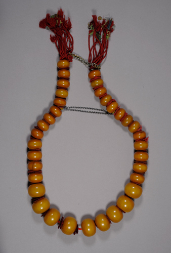 Necklace of large amber beads with red tassels on each end. The beads decrease in size at either end of the necklace and there are two metal chains connecting the sides.