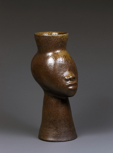 Jug with a warm brown glaze in the shape of a human face without eyes and ears and an elegant long neck.