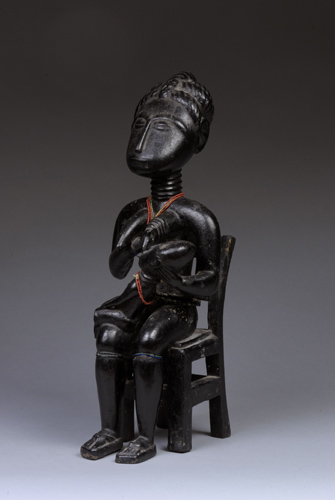 A carved wooden figure of a woman nursing a child while seated on a chair. The woman wears a red necklace.