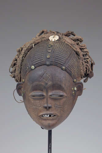 Mask of a human face with scarification patterns and an intricate textured hairstyle.