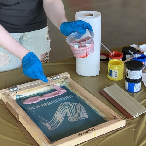 A close up shot of a student preparing a screen. The student, with blue nitrile gloves on her hands, spreads a light pink ink onto a screen.
