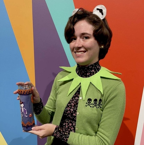 A student, in a homemade Kermit the frog costume, poses in front of the Odita mural in the Stanley lobby with their craft: a tall purple votive candle, decorated with stickers and rhinestones.