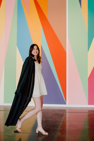 A photo of Anaka Sanders; it is an action shot of her striding in front of the lobby mural at the Stanley, dressed in her graduation robes.