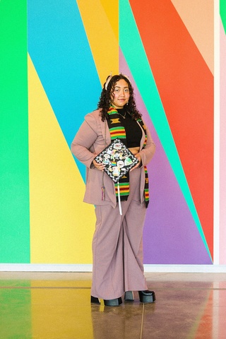 A photo of Lily Hester: they are standing in front of the mural in the Stanley's lobby, dressed up, posing with their graduation cap.