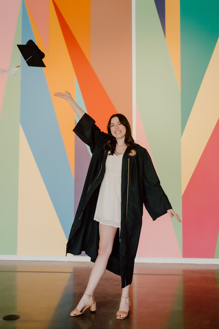 A photo of Anaka Sanders; she is posing in front of the lobby mural at the Stanley, dressed in her graduation robes, tossing her cap in the air.