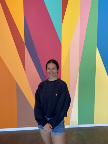 A photo of Kate Malkusak--she stands, smiling, in front of the mural in the Stanley Museum of Art lobby.