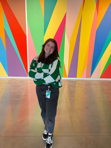 A photo of Alexis Belme: she is standing in front of the mural in the Stanley Museum of Art lobby, smiling, her arms crossed.