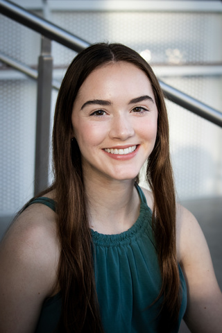 A photo of Katherine Shamdin; she is seated on stairs, smiling at the camera.