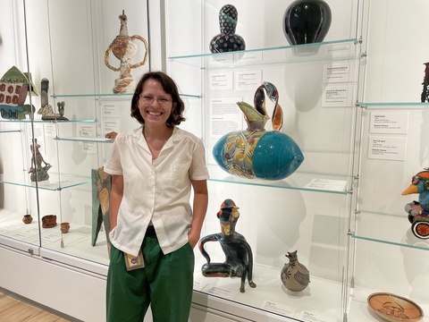 a photo of Melanie, smiling and posing in front of a glass display case of ceramics.