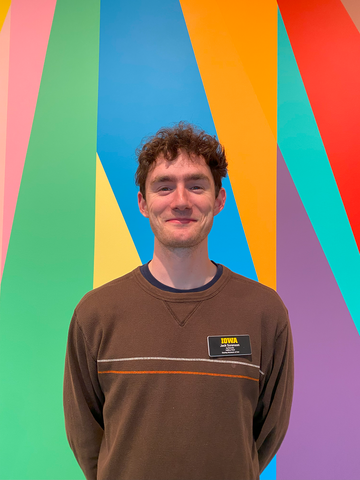A photo of gallery host Jack Sorenson. He stands, smiling, with his hands behind his back in front of the colorful lobby mural at the Stanley Museum of Art.