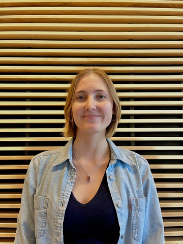 A photo of gallery host Georgia Corbin. She stands in front of the wood-slat wall in the lobby of the Stanley Museum of Art, smiling.