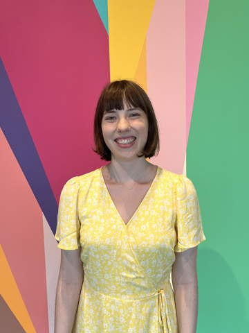A photo of Amelia, who stands smiling in front of the multi-colored mural in the Stanley Museum of Art lobby.