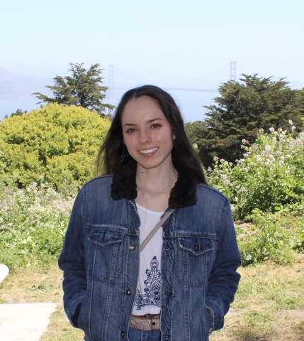 A photo of a woman with dark hair, wearing a jean jacket and smiling, with trees, green grass, and a blue sky in the background.