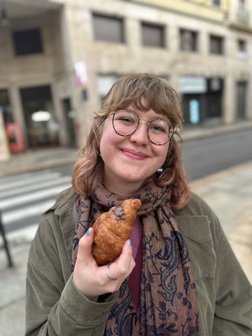 A photo of Bella taken on a street corner; she holds up a croissant in one hand, smiling.