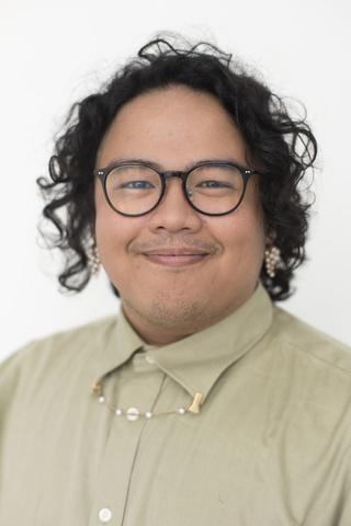 A headshot of Gallery Host Shaun Mallonga: they are wearing a light sage green button up shirt, dark brown rimmed glasses, and have dark curly hair.