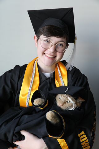 GH Jessie Meiers poses in their graduation robes with their orange cat in their arms; the cat is also dressed in graduation robes.
